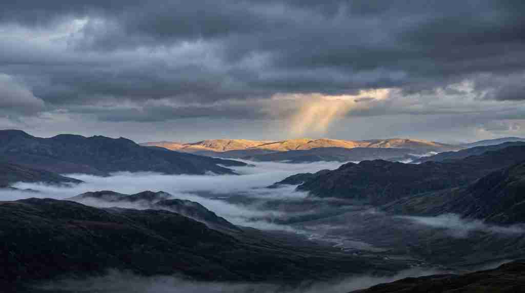 Mist fills a wide valley beneath storm clouds as a shaft of light breaks over distant ridgelines
