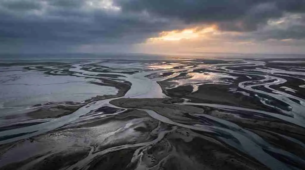 Braided river channels cross a delta plain, one channel blocked by a pale sediment bar under stormy light