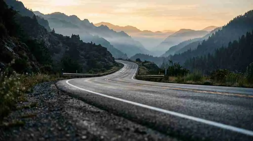 Eine zweispurige Bergstraße, die sich im Morgengrauen durch den Nebel schlängelt, mit warmem Licht am Horizont