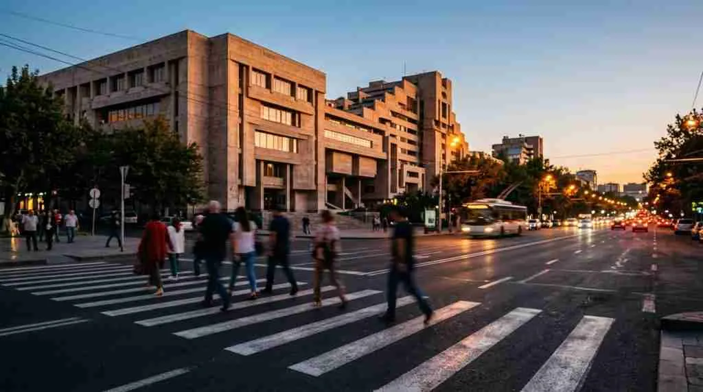 Verschwommene Fußgänger überqueren in der Abenddämmerung eine breite Allee neben einem sowjetisch-modernistischen Betongebäude in Jerewan.